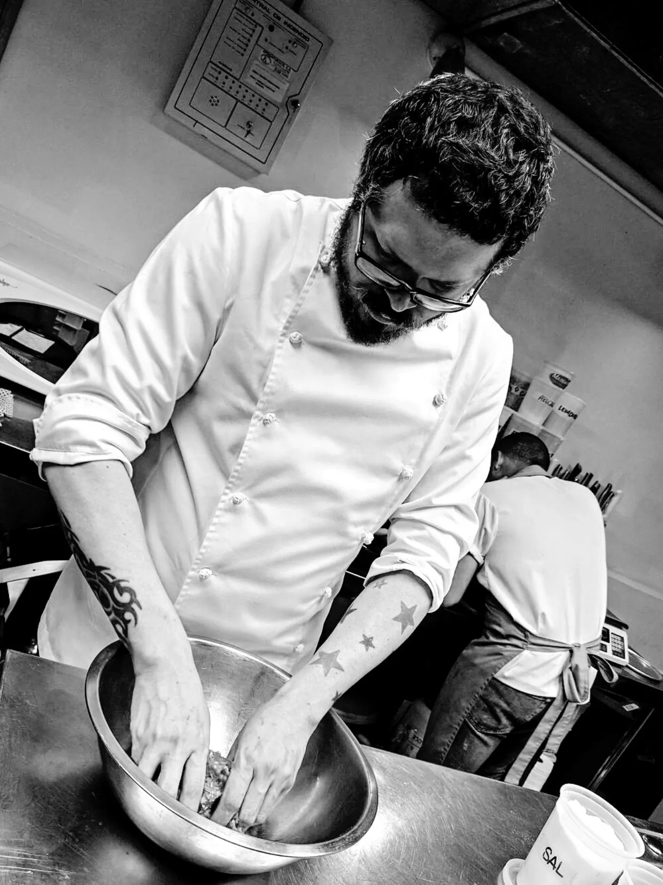 Chef con chaquetilla blanca amasando ingredientes en un bol metálico en plena cocina profesional. Blanco y negro.