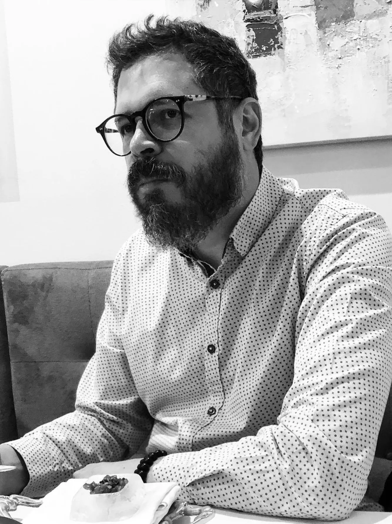 Chef John Guerrero, con barba y gafas redondas, sentado a una mesa con un plato emplatado. Retrato en blanco y negro.